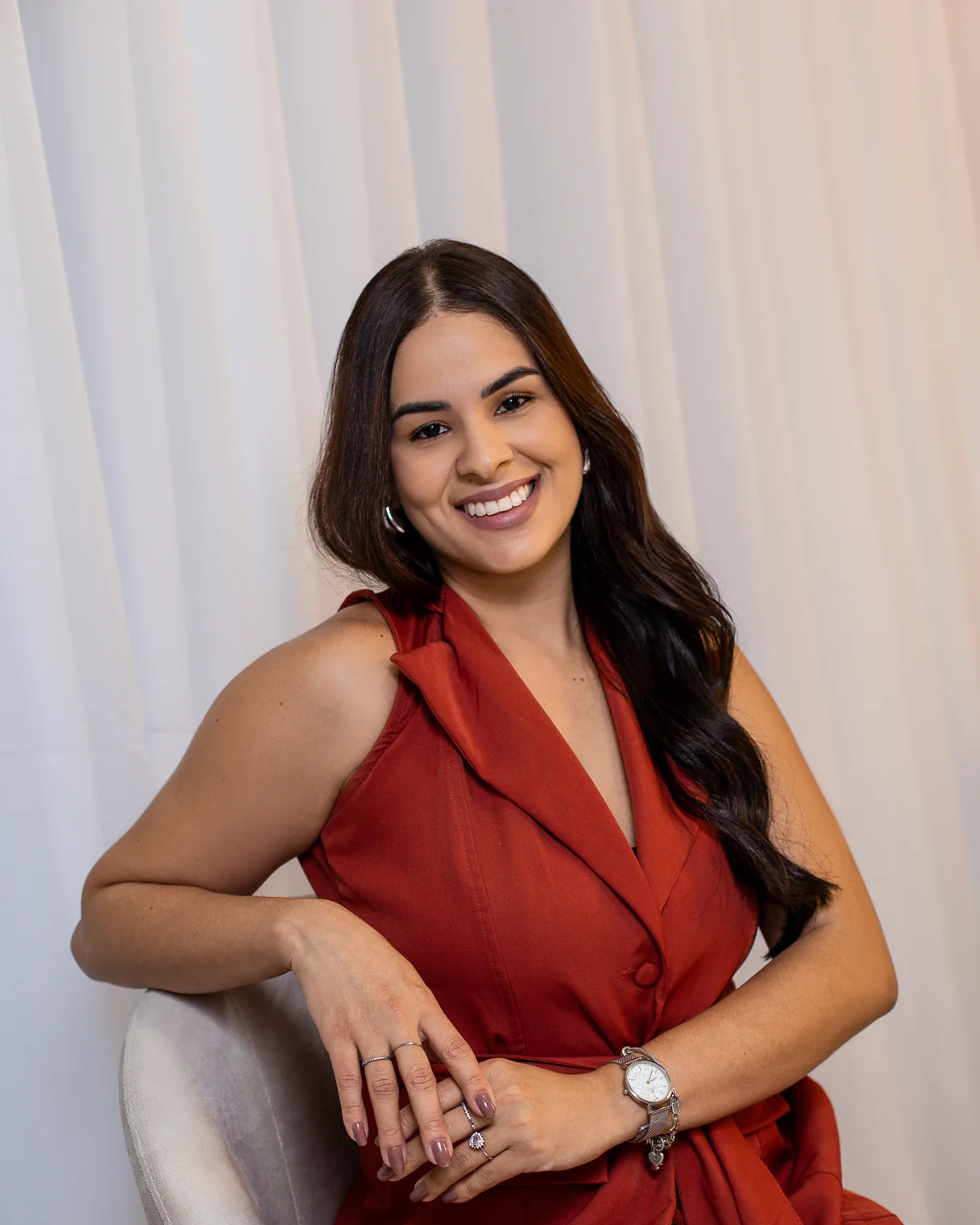 Mislley Santos sentada em uma poltrona clara, de colete vermelho, sorrindo para a câmera.