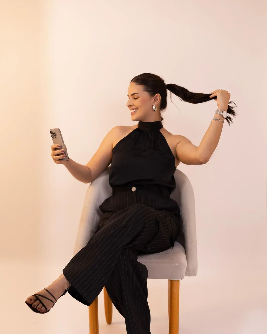 Mislley Santos sentada em uma poltrona, rindo, olhando para o celular em uma das mãos e segurando o rabo de cavalo com a outra.
